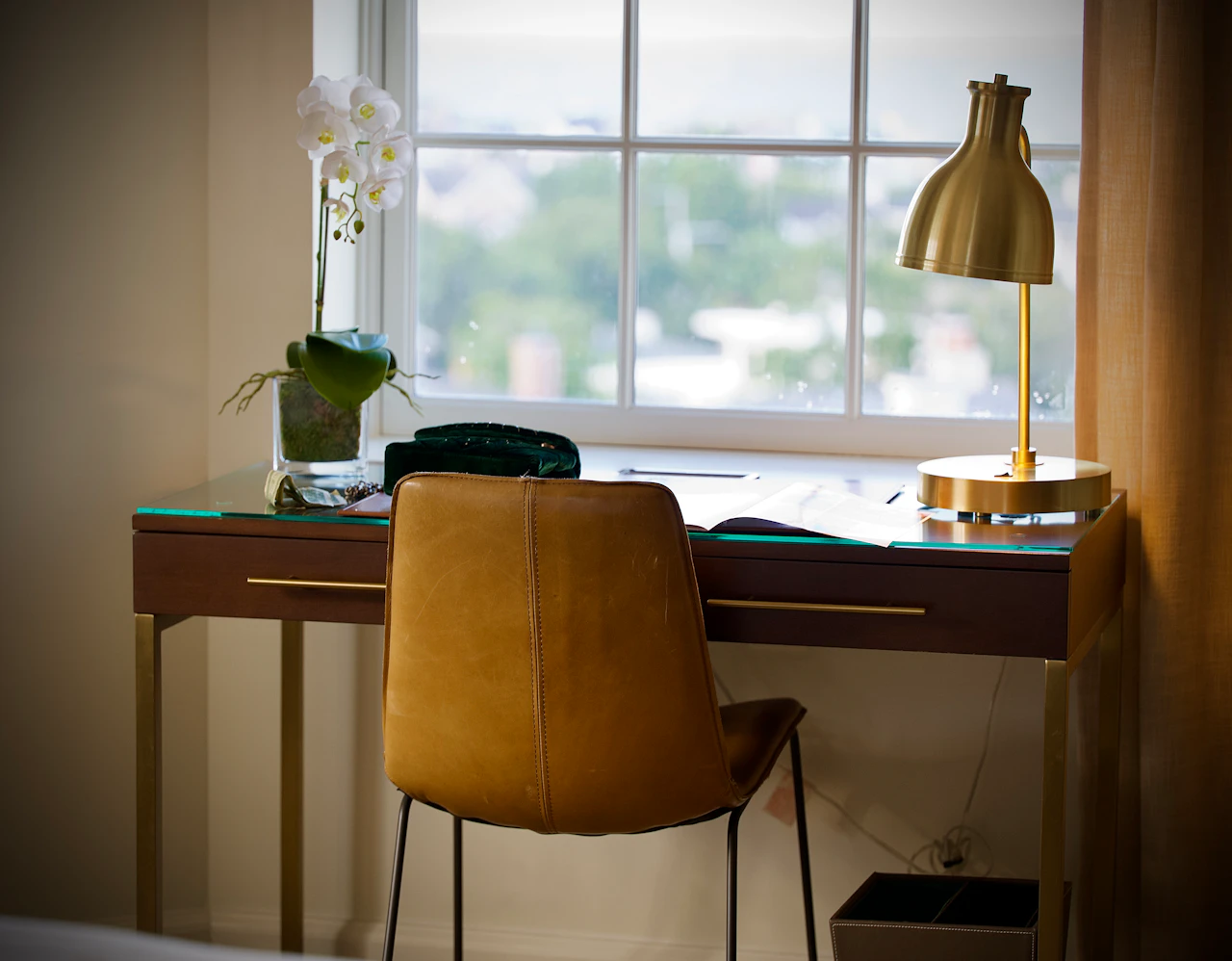A hotel room desk with a brass lamp, leather chair, and a window onto green hills