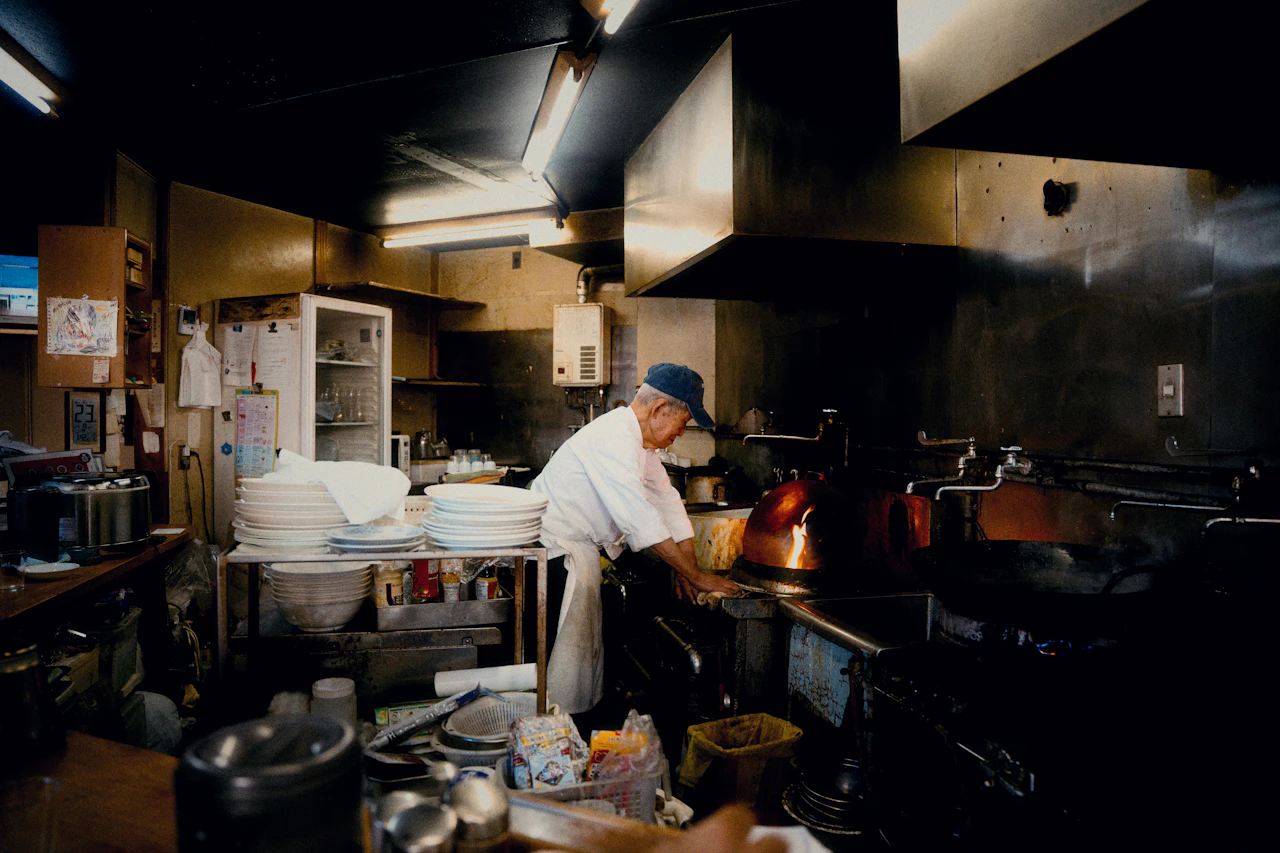 Small restaurant owner at a wooden counter working through end-of-day numbers with a ledger and calculator, warm evening light